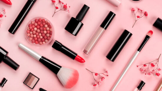 Various cosmetic accessories for makeup and manicure on trendy pastel pink background with red flowers. Blush, brush, eye shadow, mascara, perfume, lipstick, nail Polish. Skin care products.